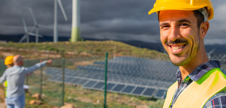 L'image montre une personne portant un casque de sécurité jaune et un gilet de sécurité réfléchissant se tenant au premier plan. En arrière-plan, il y a des éoliennes et des panneaux solaires installés sur un champ. Une autre personne est visible en arrière-plan, pointant vers les éoliennes. Le ciel est nuageux et le cadre semble être un site extérieur dédié aux énergies renouvelables.